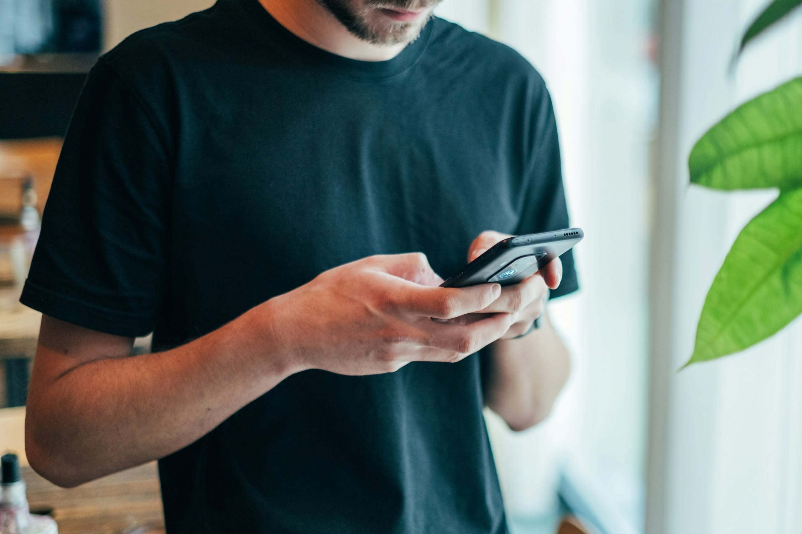 Close-up of a person in a black T‑shirt holding a smartphone with both hands, looking down at the screen near a bright window; a green plant leaf is visible at the edge.