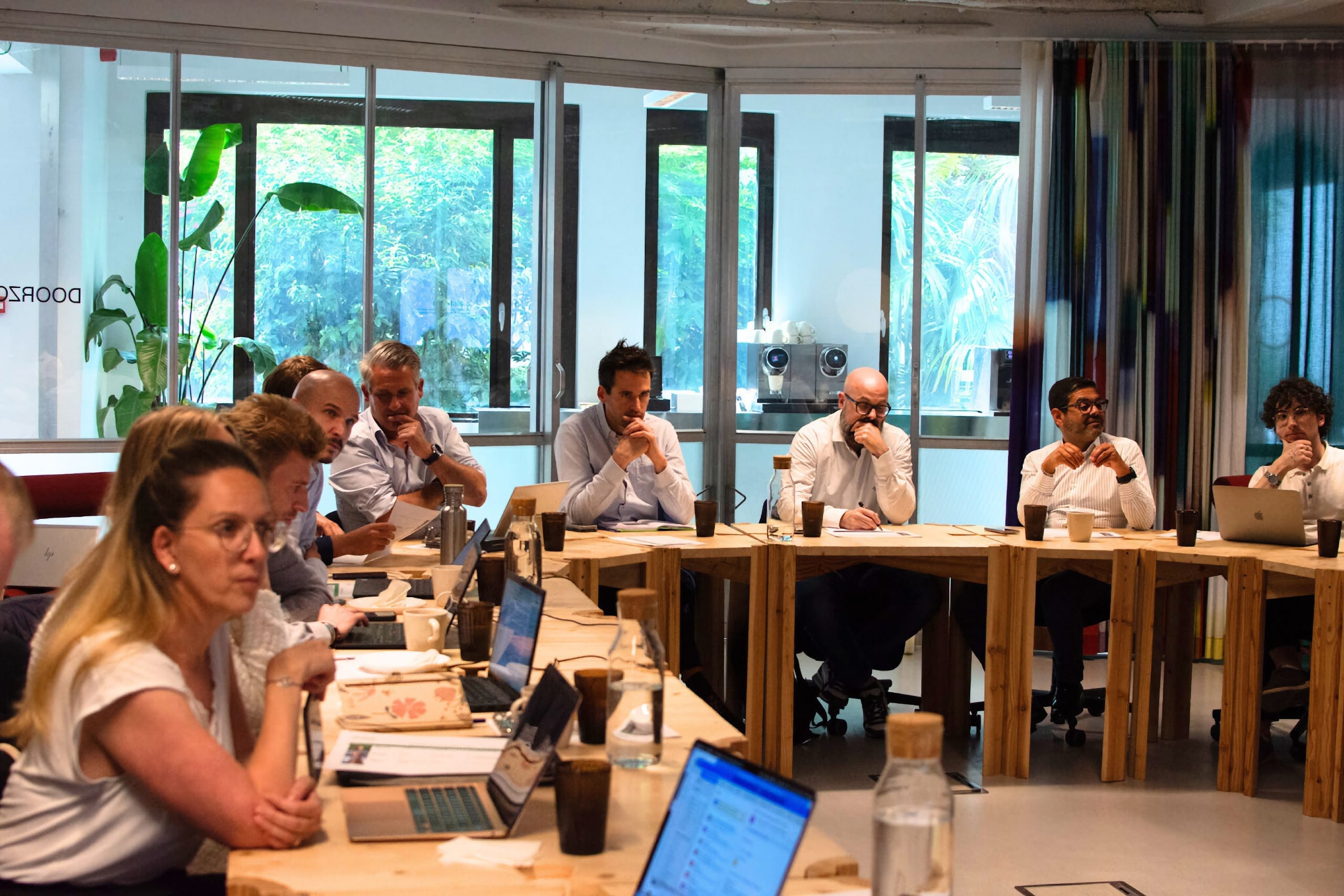 A diverse group of people sit around a U‑shaped table in a bright meeting room, focused on discussion with laptops, notebooks, coffee cups, and water bottles in front of them; large windows show leafy greenery outside and colorful curtains line one wall.