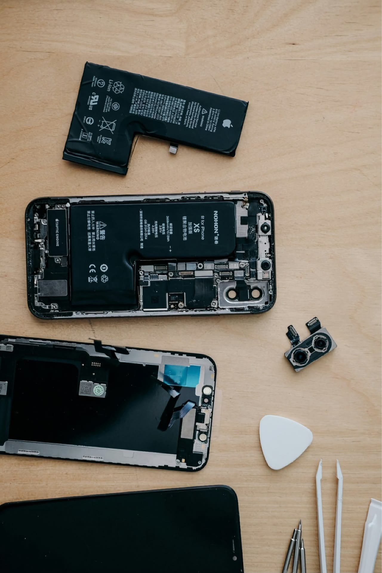 Disassembled smartphone parts on a wooden desk: open phone with battery and motherboard visible, loose rear camera module, separate screen assembly, spare battery, plastic opening pick, and white pry tools