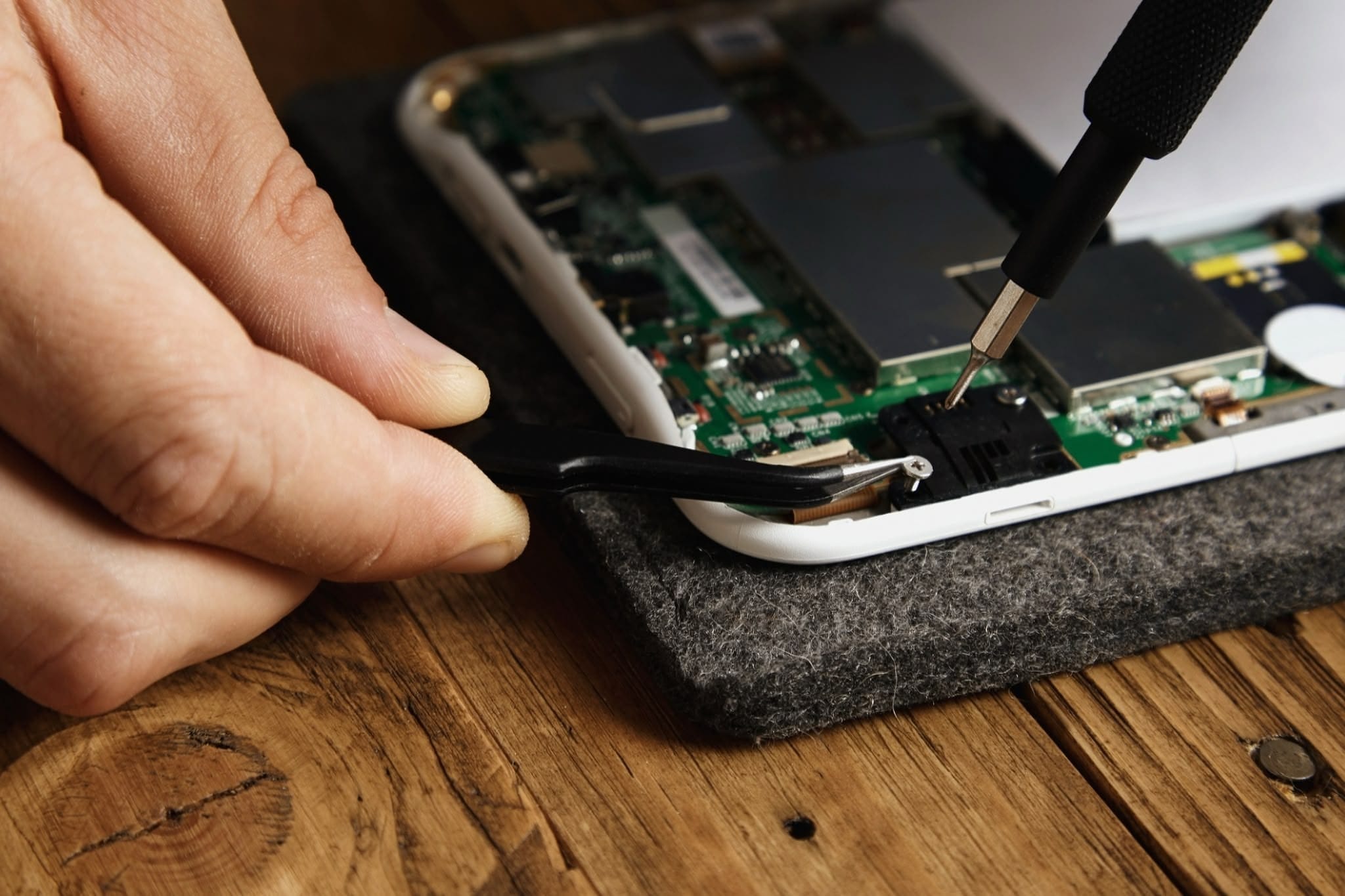 Close-up of hands repairing a smartphone on a felt pad: one hand holds angled tweezers near a connector while a precision screwdriver adjusts a small screw on the exposed circuit board; wooden table surface visible.