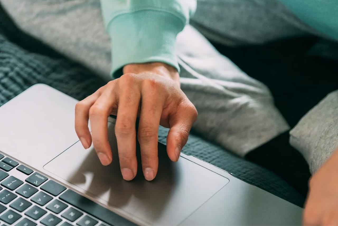 Close-up of a hand using a laptop trackpad while seated, with soft fabric clothing visible; keyboard edge is partially in frame.