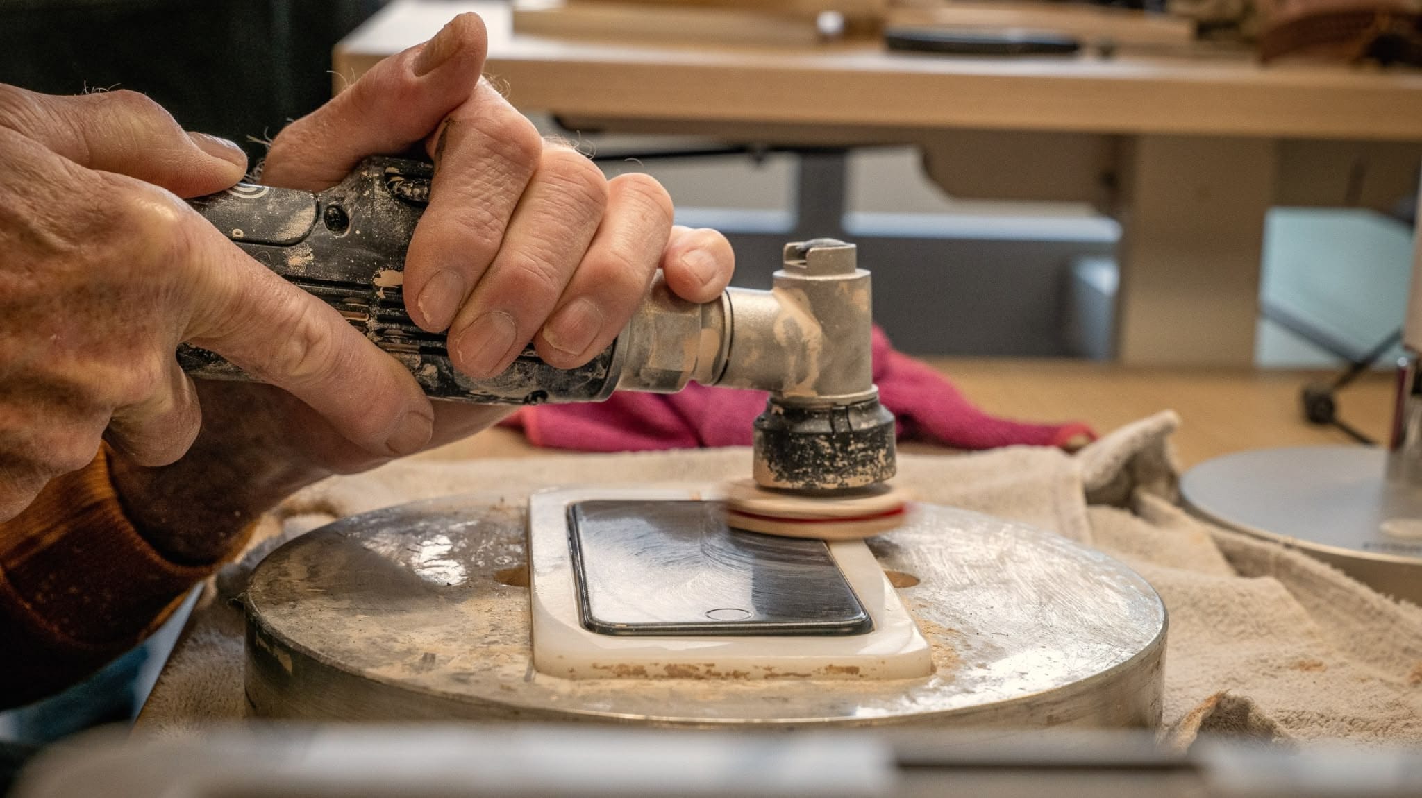 Close-up of hands using a rotary polishing tool to buff a smartphone screen mounted on a jig, with dust on the tool and a towel beneath the setup.