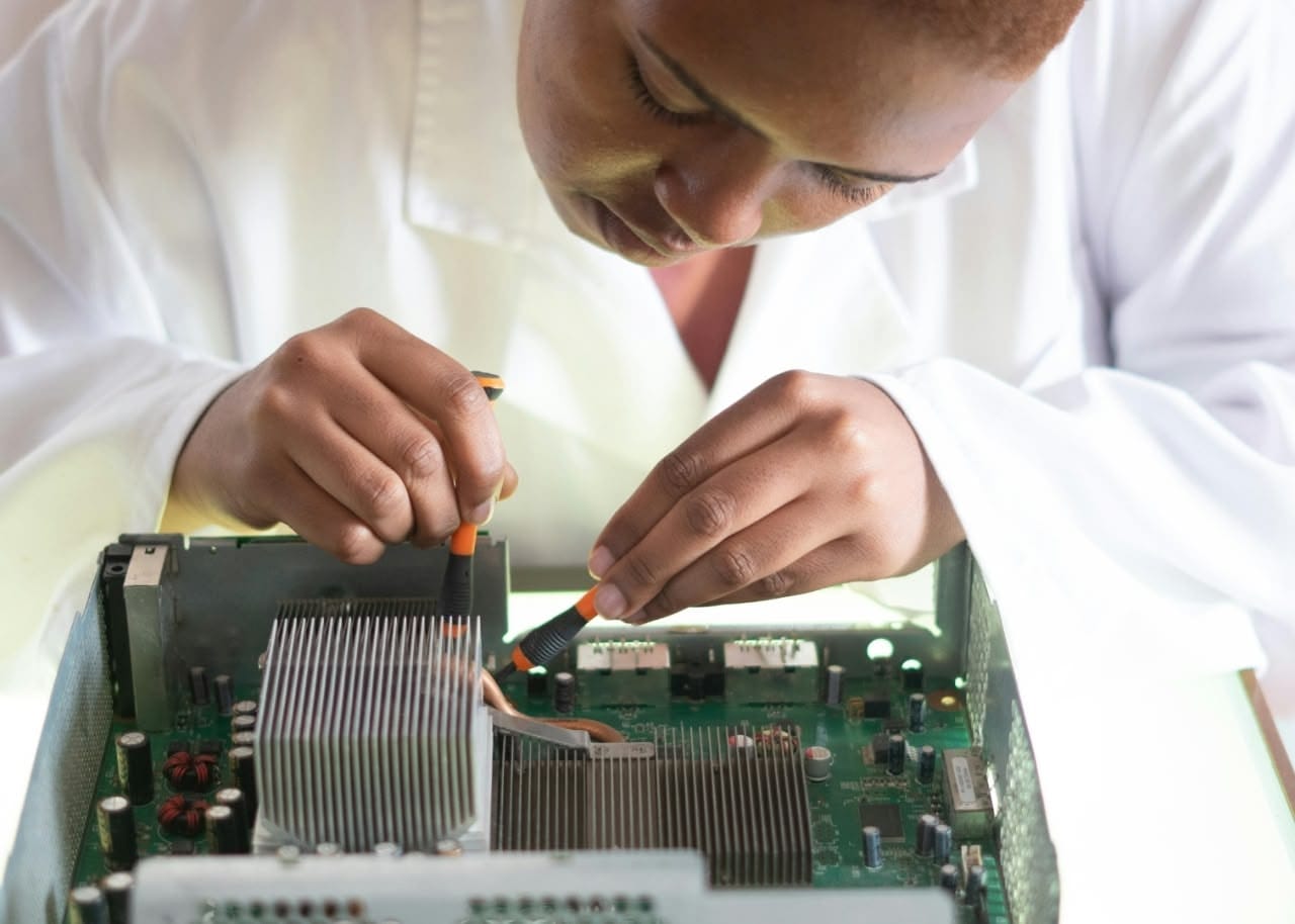 Person in a white lab coat concentrates while using two screwdrivers to work on a large circuit board with a metal heatsink inside an open device chassis.